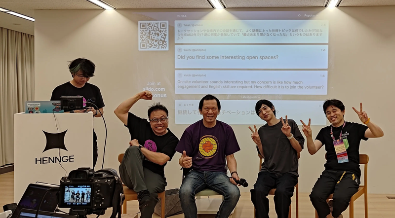 Photograph of four men posing on a stage with a projection screen showing "Q&A" and "slido.com" and a "HENNGE" podium.